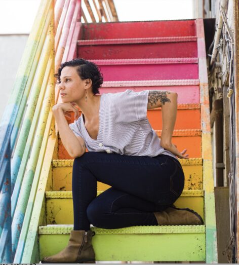 Cara sits on a multicolored staircase in a white shirt, blue jeans, and brown boots. Cara Has cropped, curly black hair and brown skin. Photo courtesy of the artist.
